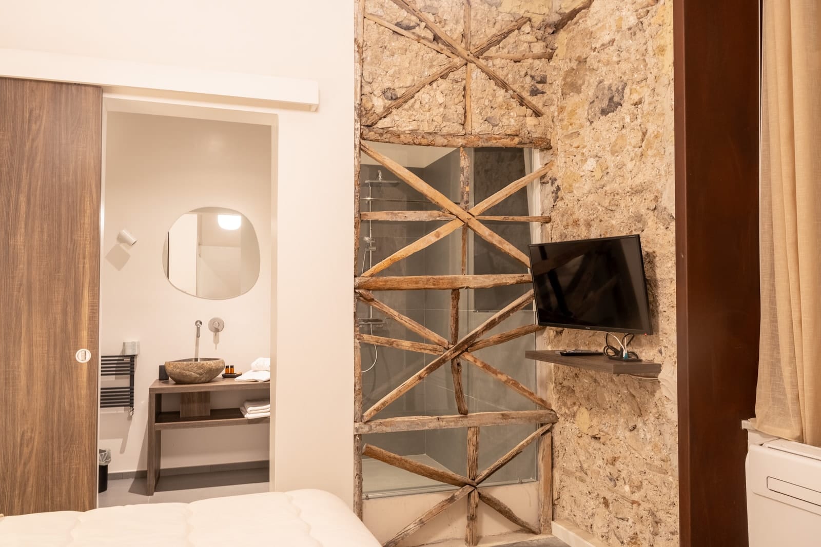 La camera Zeus: la porta scorrevole aperta sul bagno, i controventi di legno e il televisore sulla parete di pietra